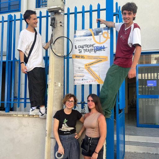 Group of four young people posing in front of a poster promoting a youth center project, with two individuals standing on a fence and two on the ground, smiling.